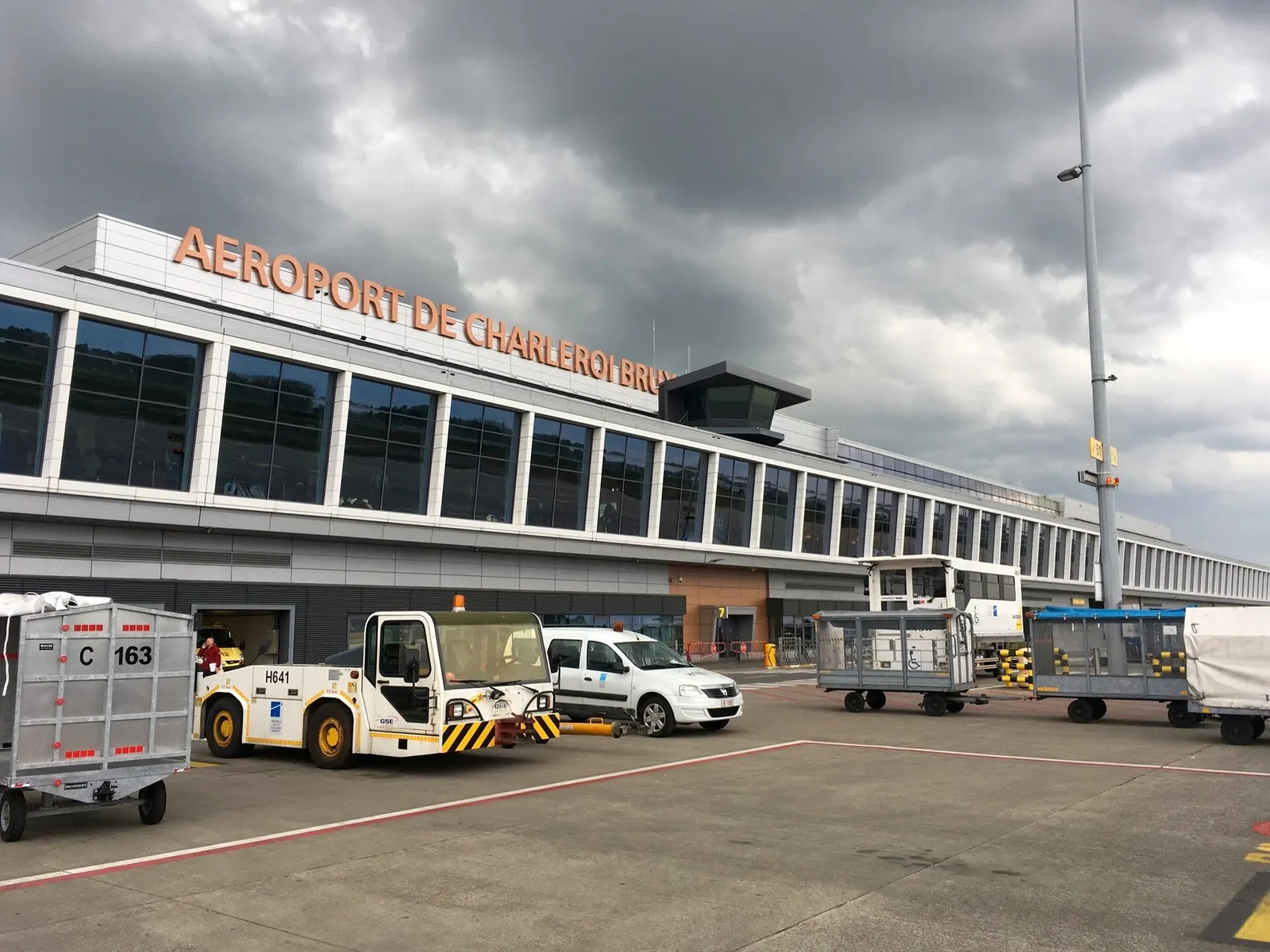 Airside façade of Charleroi Terminal 1 with AEROPORT DE CHARLEROI BRUXELLES SUD lettering and ground-support vehicles on the apron.
