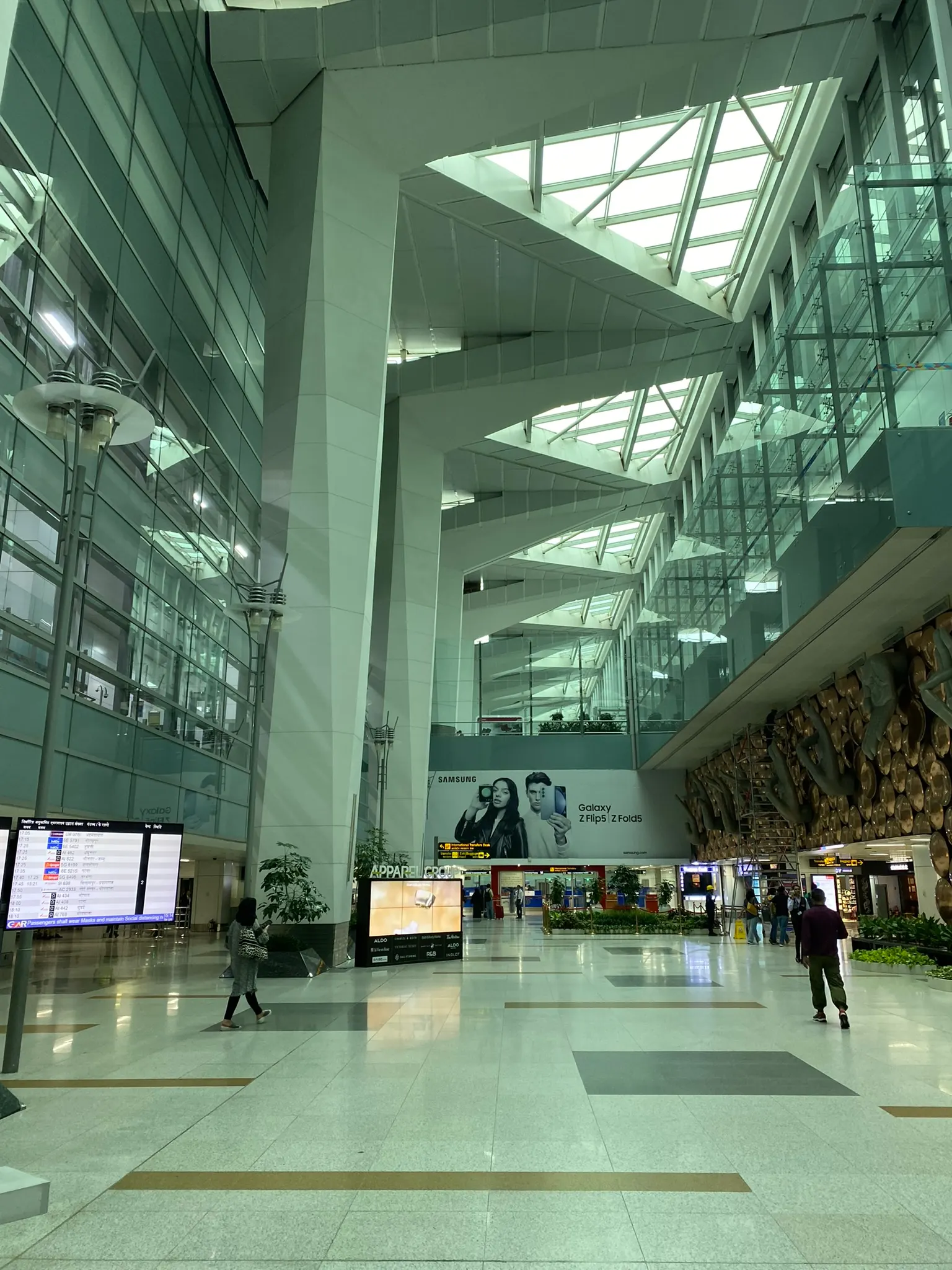 Travelers walking through a bright, modern interior corridor with shops and gate signage inside Indira Gandhi International Airport.