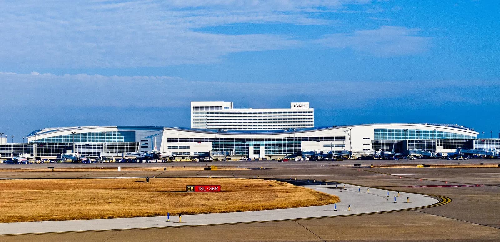 Wide exterior view of DFW Terminal D's curved glass facade with the Grand Hyatt hotel rising behind it under a cloudy sky.