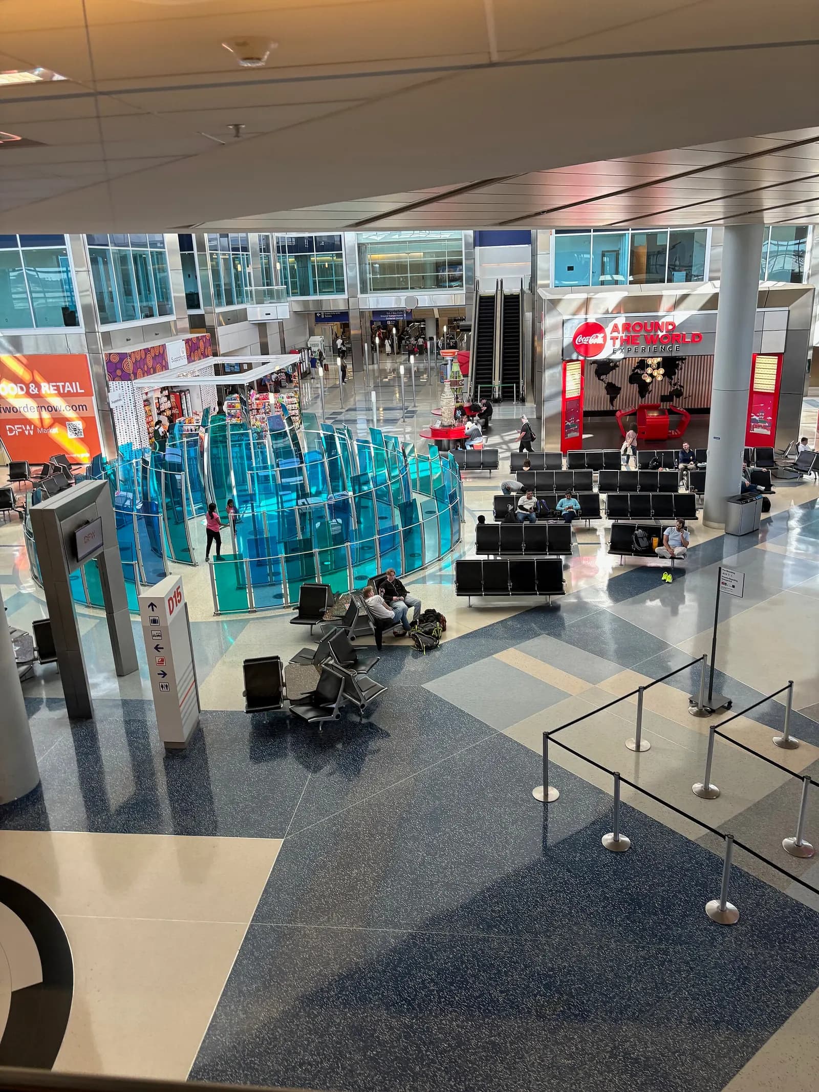 Bright Terminal D concourse at DFW with travelers walking past shops, a large sculpture, and an elevated international arrivals catwalk in the background.
