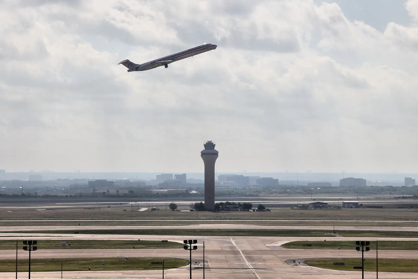 DFW's tall air traffic control tower standing against a blue sky with part of an aircraft tail visible at the edge of the frame.