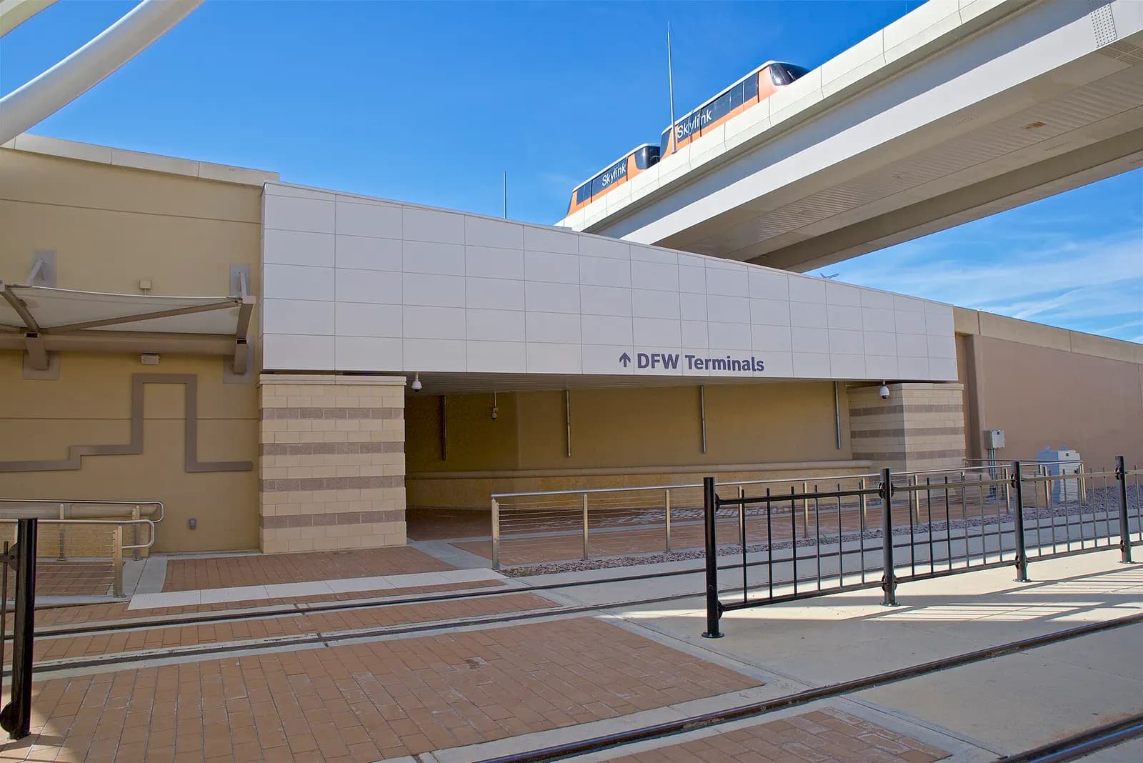 Pedestrian walkway and glass entrance to DFW Terminal B at the TEXRail station, with the elevated Skylink track visible overhead.