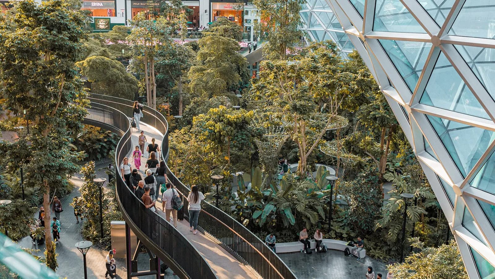A walking path through The Orchard, a lush indoor tropical garden at Hamad International Airport, with travelers passing beneath a modern glass ceiling.