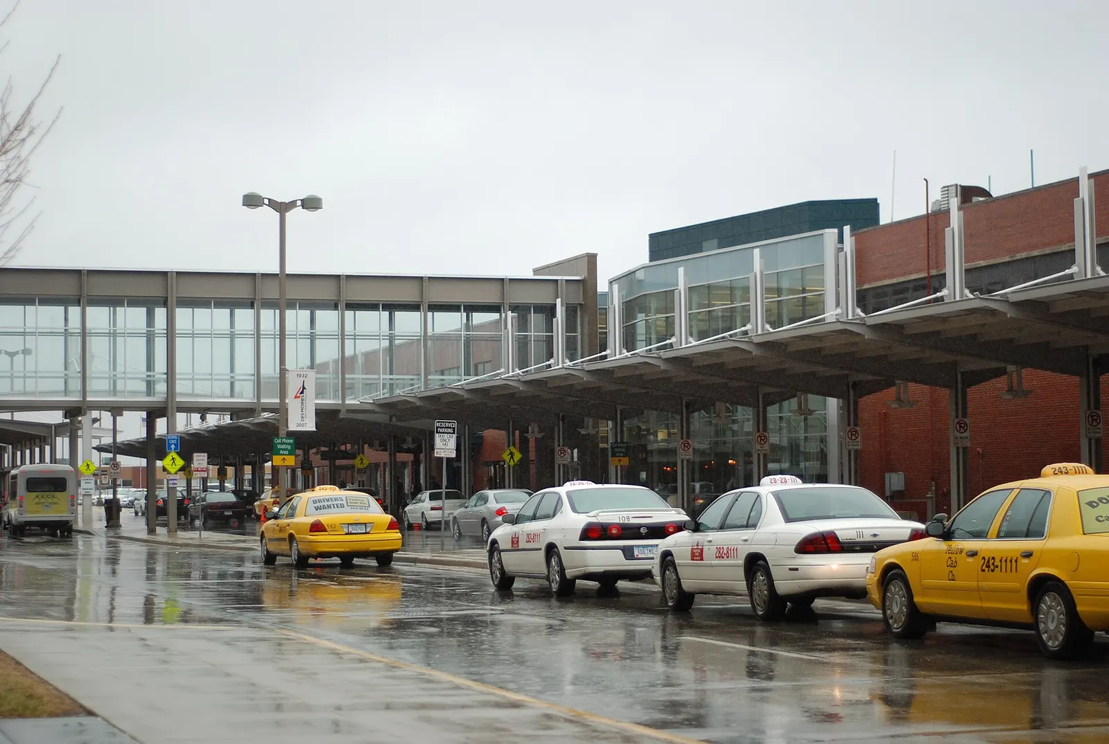 Landside curb and skybridge at Des Moines International Airport on a wet day, with taxis waiting.
