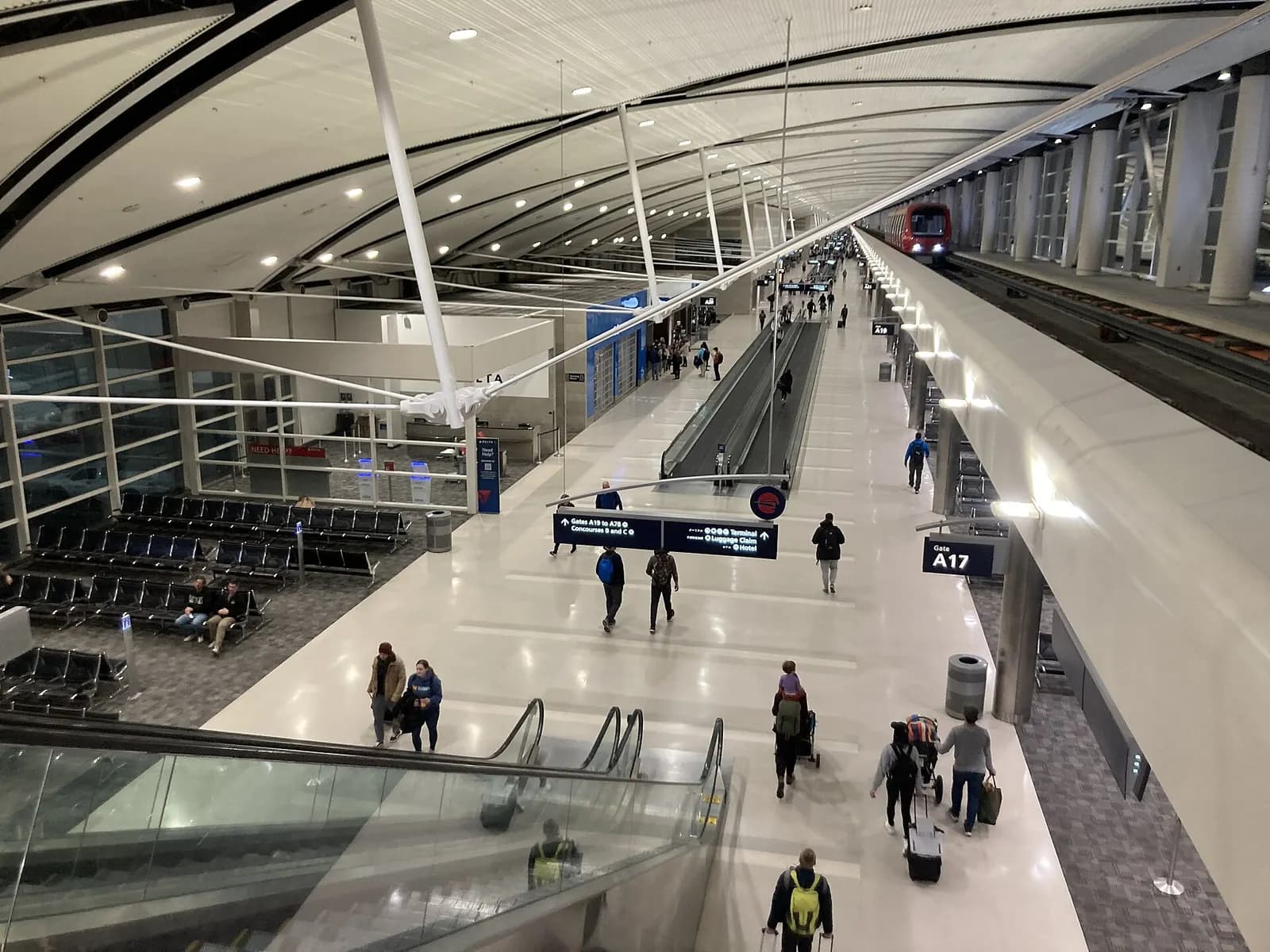 Travelers walk along a bright, wide Concourse A corridor with gate seating areas and large windows at Detroit Metropolitan Airport.