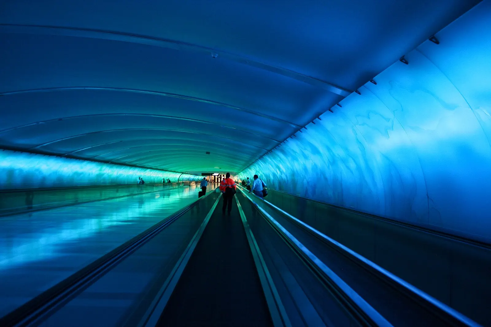 A long underground pedestrian walkway lined with illuminated glass panels in purple and blue, connecting terminals at Detroit Metro Airport.