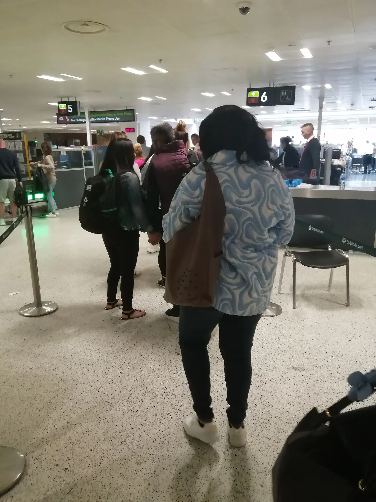 Passengers queue at the security screening lanes inside Dublin Airport, with belts, bins, and metal-detector arches visible.