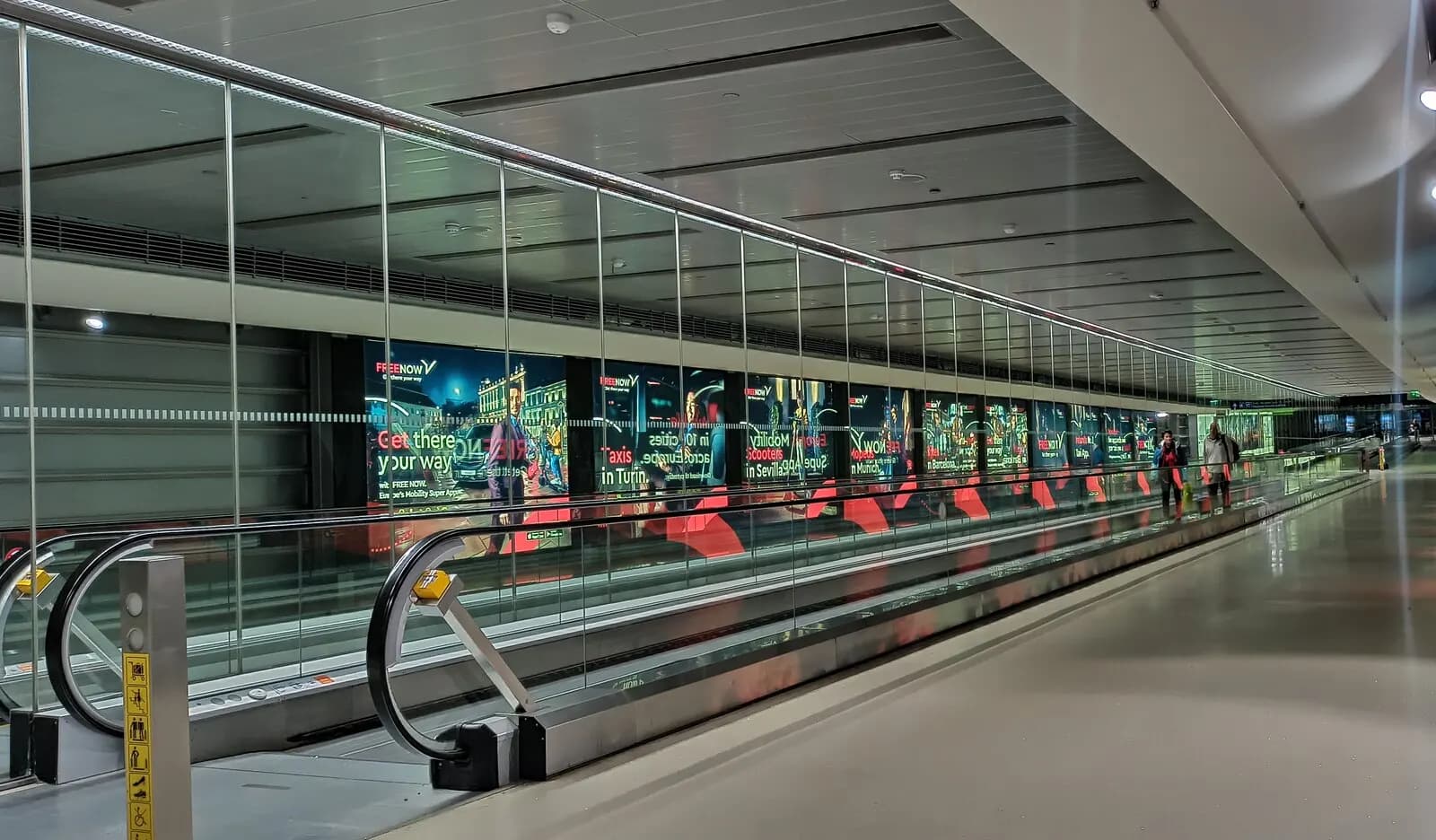 A long, brightly lit passenger corridor inside Dublin Airport with moving walkways and glass walls.