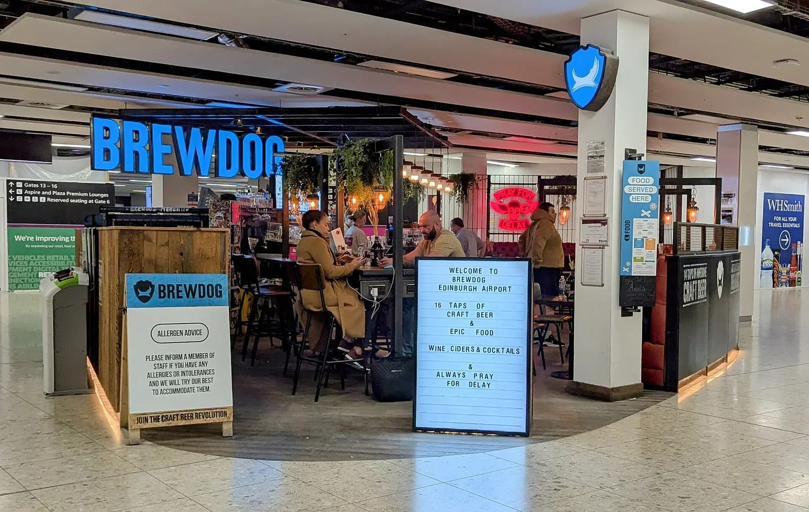 The interior of a BrewDog bar at Edinburgh Airport with wooden tables, pendant lighting, and the bar counter along the back wall.