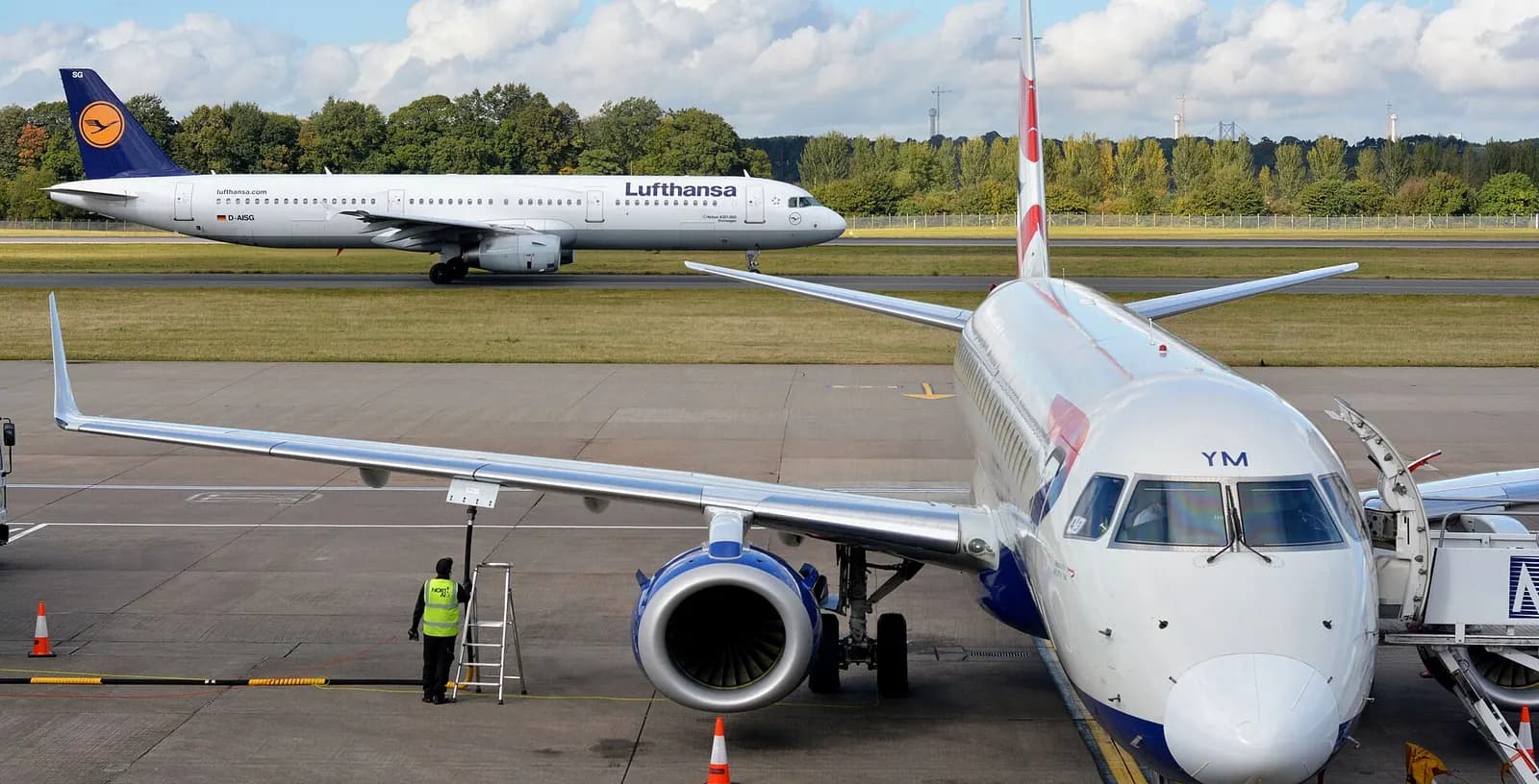 A wide view of the Edinburgh Airport apron with a British Airways regional jet at a stand and a Lufthansa Airbus taxiing past the terminal.