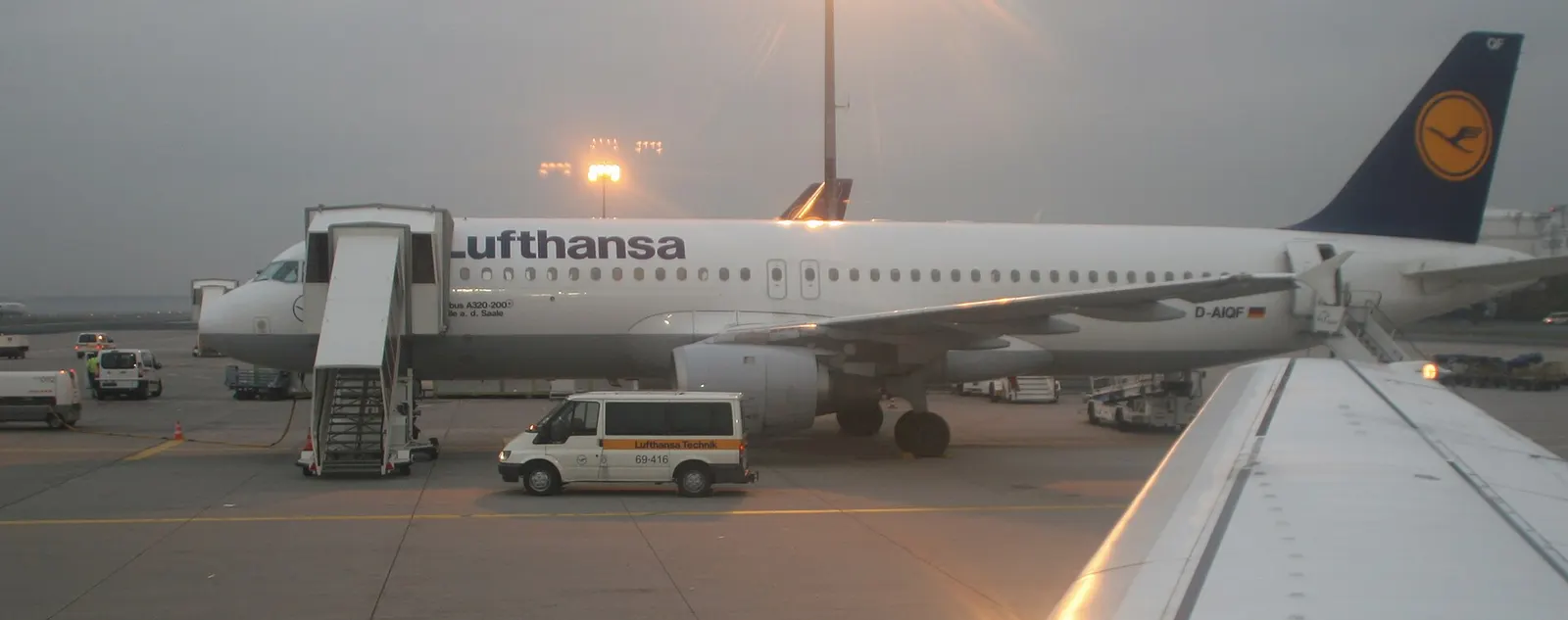 A Lufthansa Airbus A320 parked at a remote stand on the tarmac, reached from the terminal by airport shuttle bus.