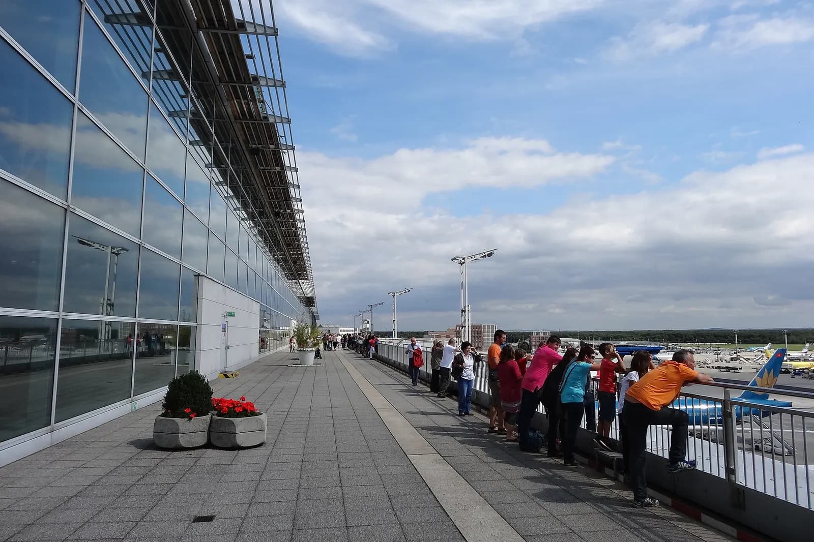 Visitors on the open-air observation terrace at Terminal 2 overlooking the ramp, with aircraft and the terminal facade in the evening light.