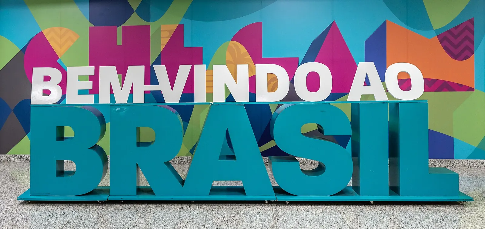 Large 'Bienvenido Brasil' welcome sign displayed in a Galeão terminal arrival area with travelers nearby.
