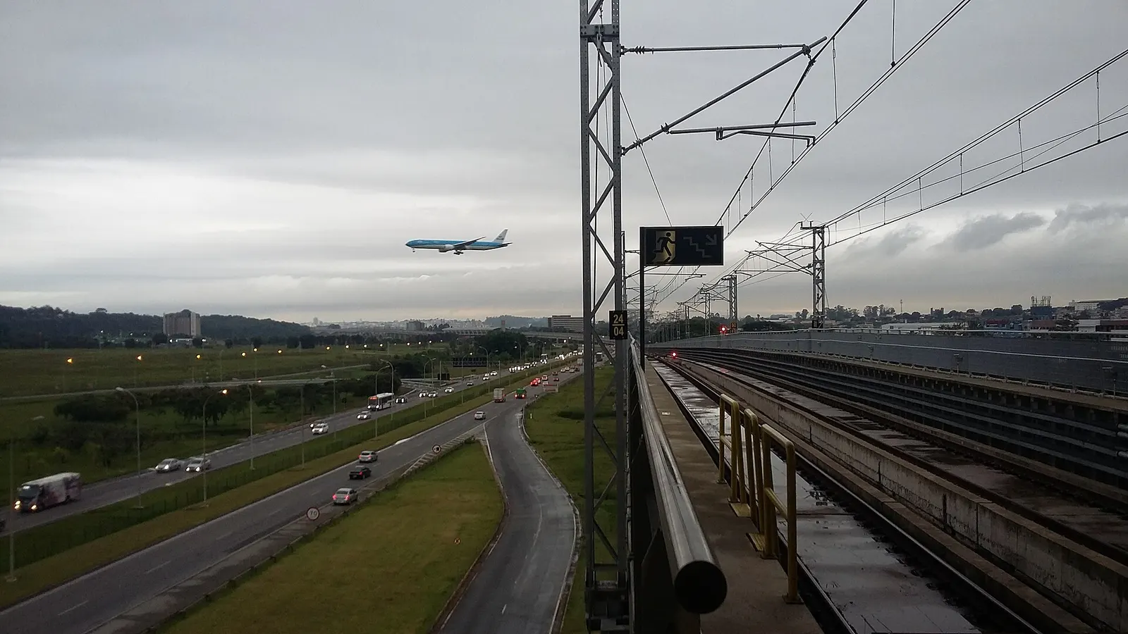View from Aeroporto Guarulhos station showing CPTM Line 13-Jade tracks beside the Hélio Schmidt highway and airport ramps with a KLM aircraft.