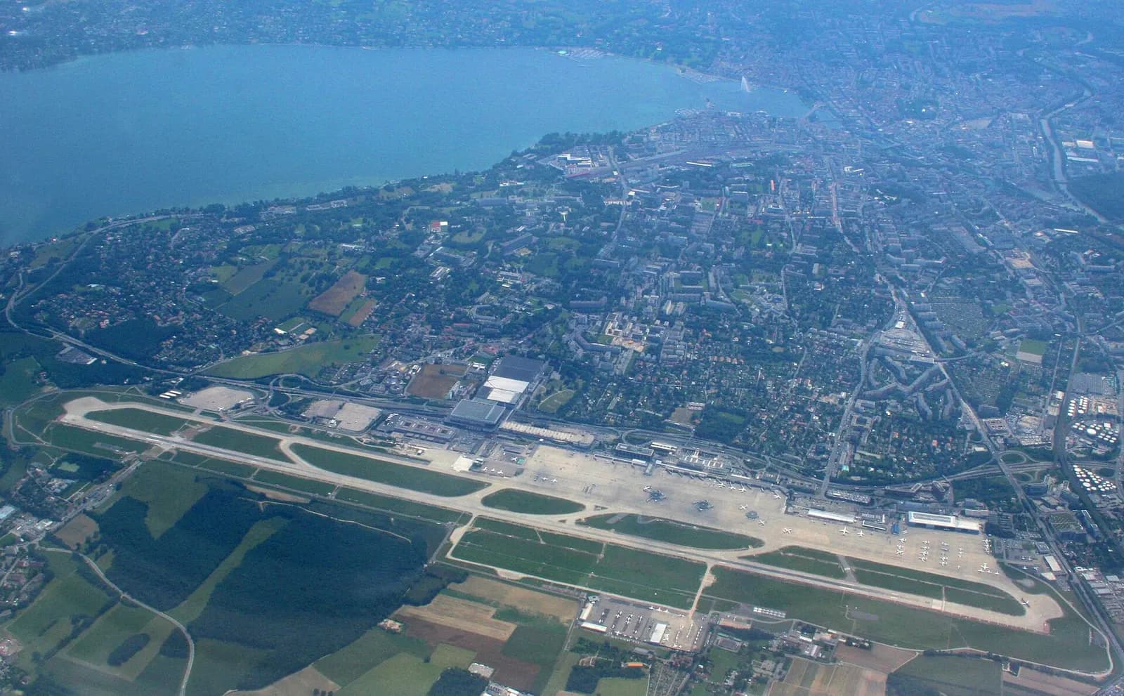 An aerial view of Geneva International Airport's runways and terminal buildings with Lake Geneva and the city skyline in the background.