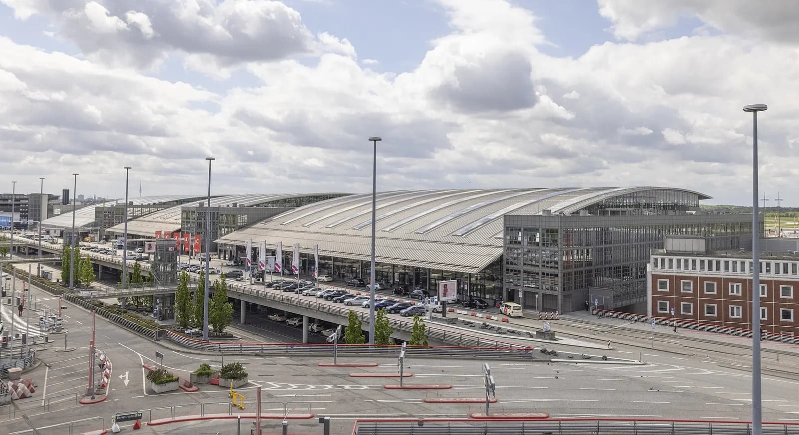 A wide daytime view of Hamburg Airport's modern terminal buildings with glass facades, jet bridges, and apron activity.