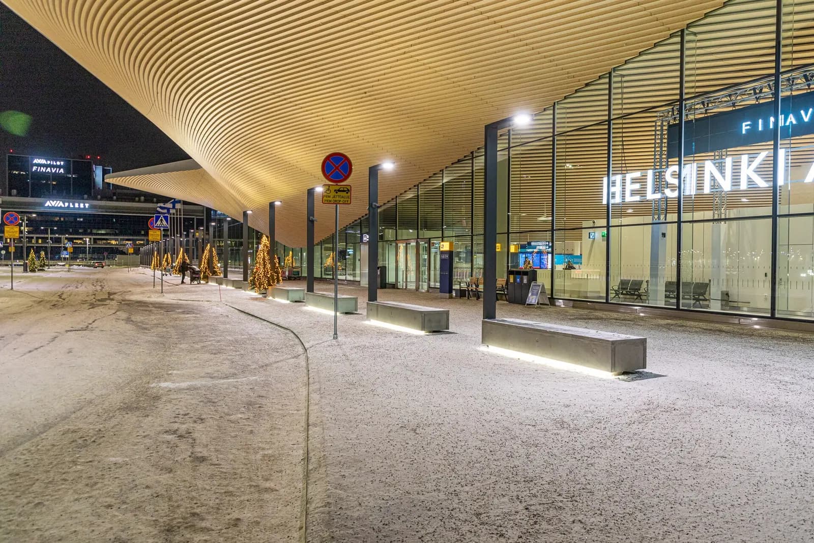 The glass-fronted entrance to Helsinki Airport's new departure hall with travelers and luggage carts outside.