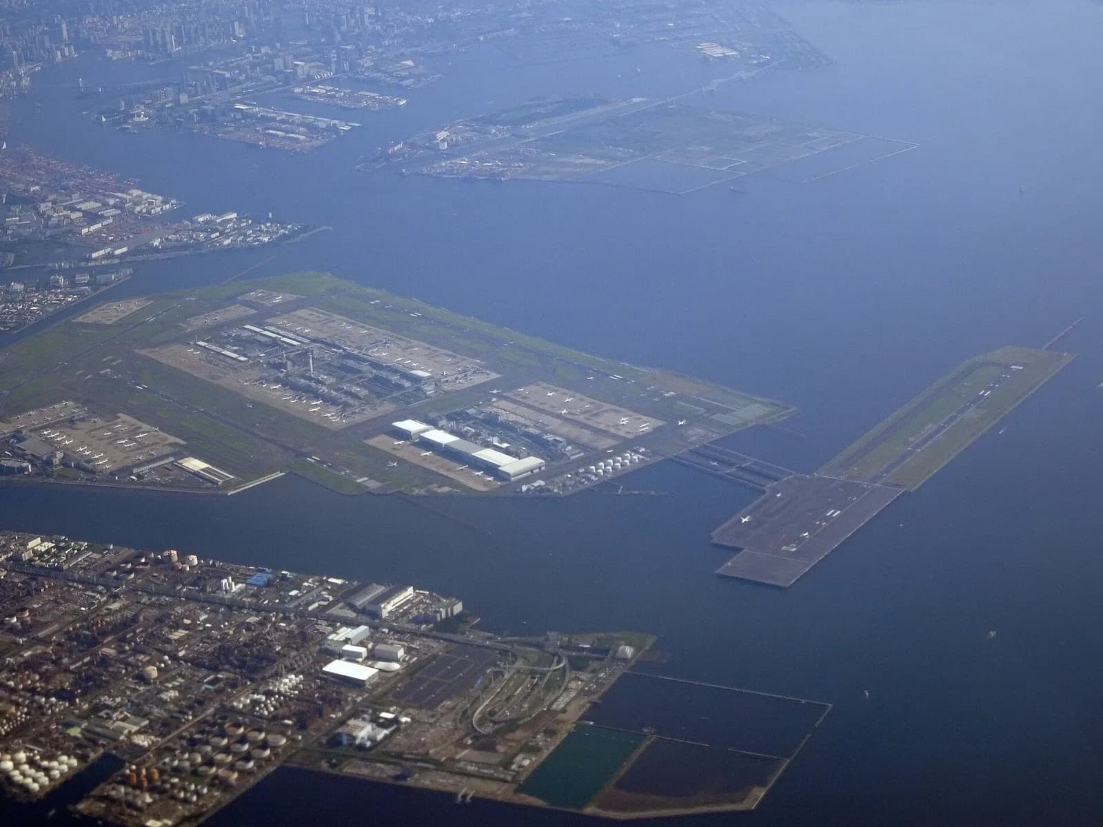 Aerial view of Tokyo Haneda Airport on a peninsula in Tokyo Bay, showing multiple runways, terminal buildings, and surrounding water.