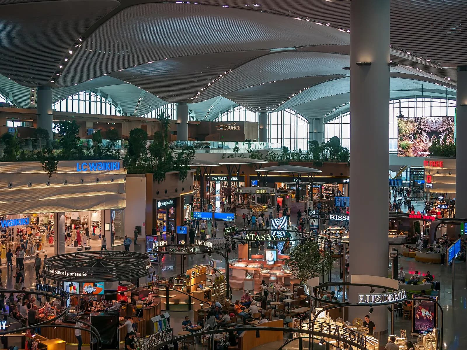 Bright duty-free shopping concourse inside Istanbul Airport viewed from a vestibule, with retail storefronts lining the walkway.