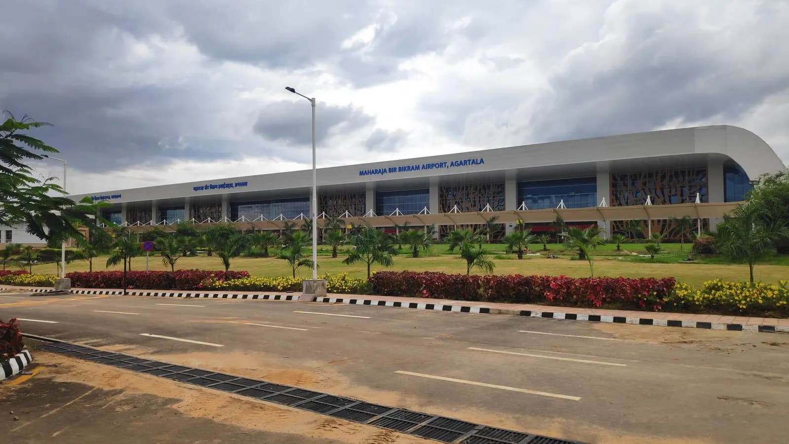 Daytime landside view of the Maharaja Bir Bikram Airport terminal facade in Agartala, with bilingual MAHARAJA BIR BIKRAM AIRPORT AGARTALA signage and tree-motif columns.