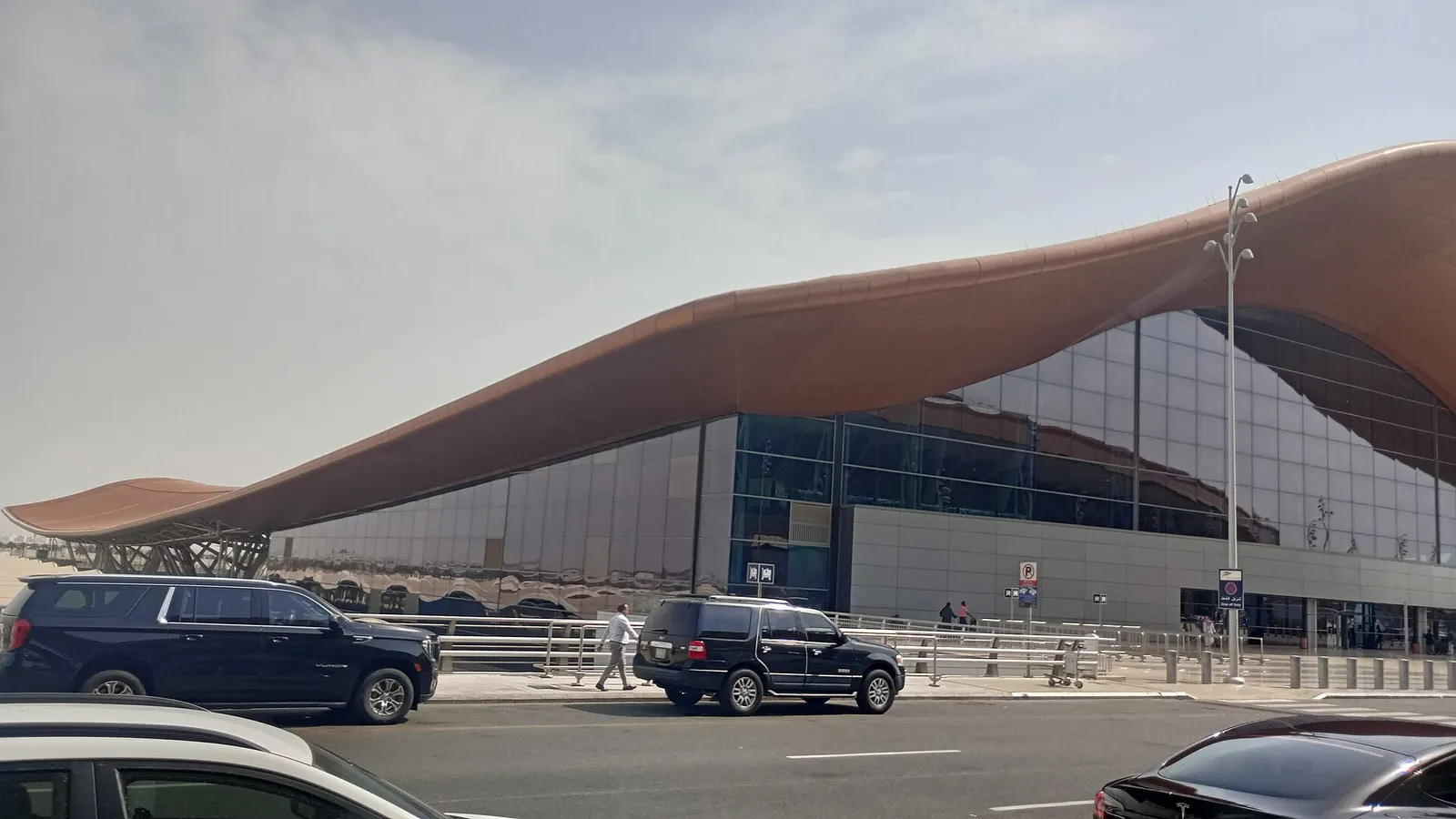 The exterior of a modern Jeddah airport passenger terminal rises above the approach road on a clear day.