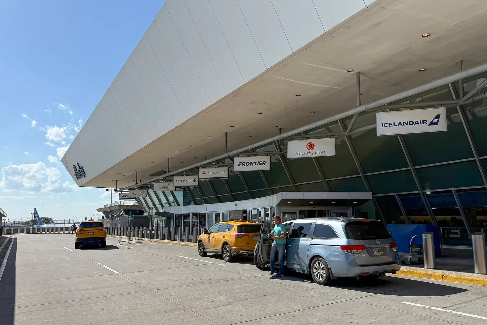 The departures-level roadway and facade of Terminal 7 show curbside drop-off lanes, terminal signage, and the building's glass-fronted entrance.