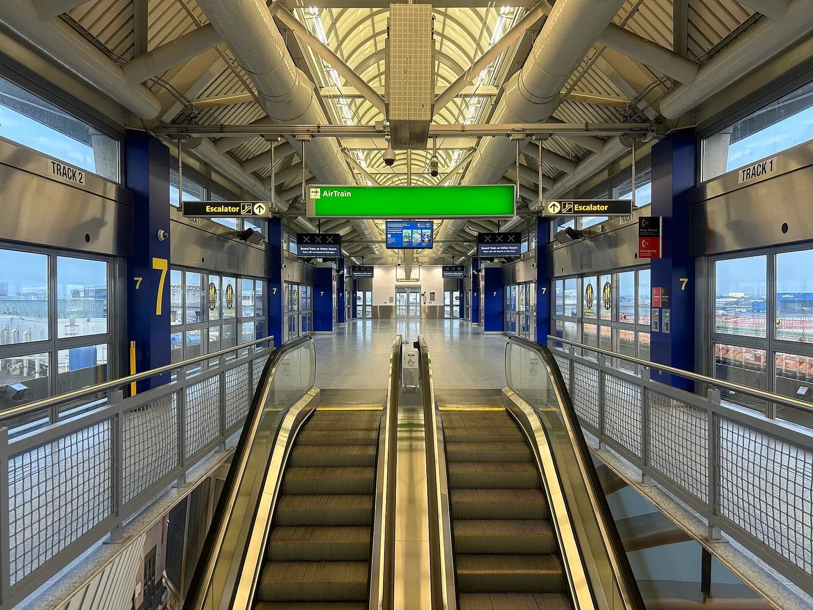 The elevated AirTrain JFK station at Terminal 7 has glass-enclosed platforms, signage, and tracks running above the roadway below.