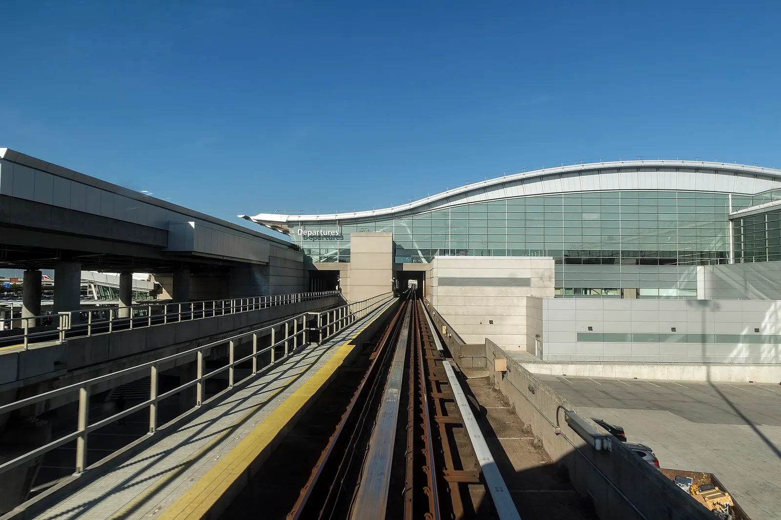 An elevated AirTrain track curves toward Terminal 4, with the guideway, support columns, and station structure rising above the terminal access roads.