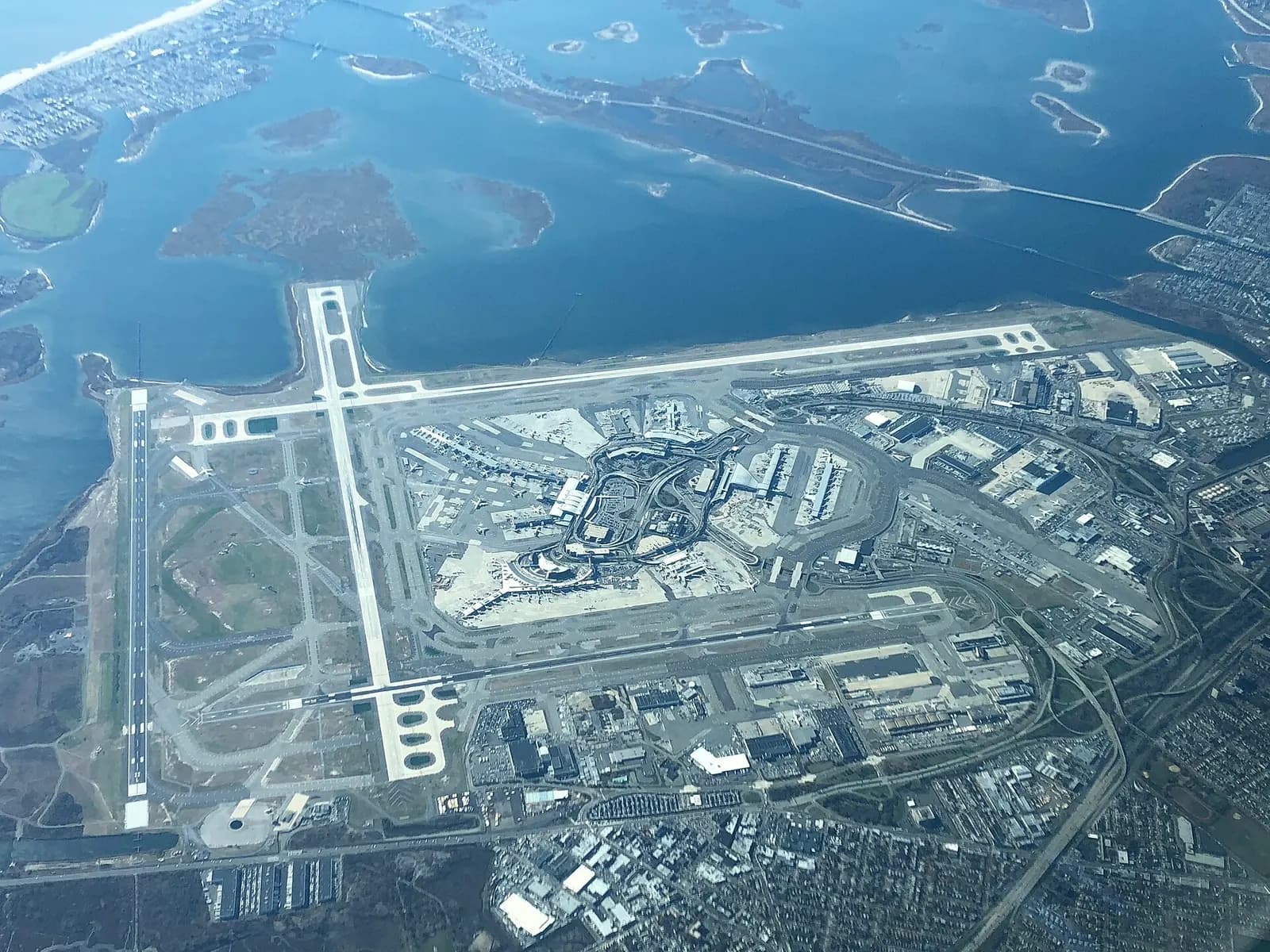 An aerial view shows the sprawling layout of John F. Kennedy International Airport with multiple terminals, runways, taxiways, and surrounding roads in Queens.