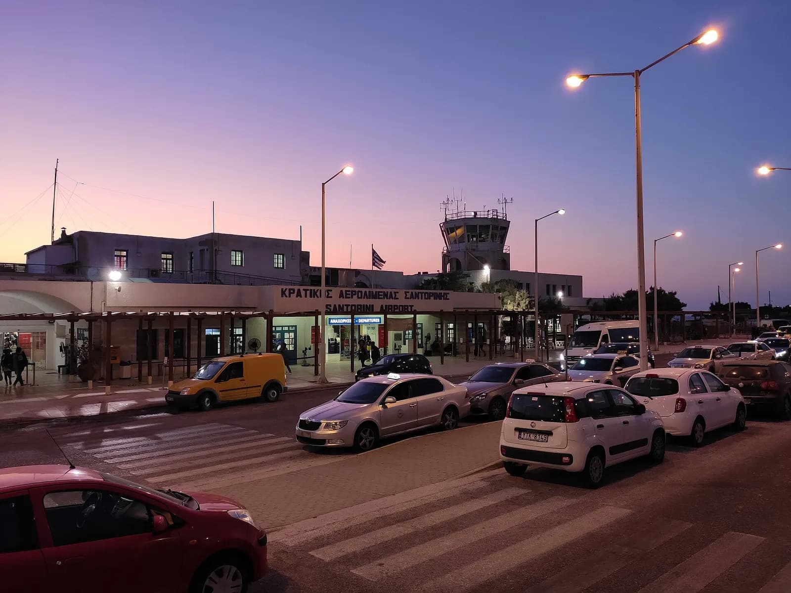Santorini Airport terminal at dusk with SANTORINI AIRPORT lettering, taxis, and control tower