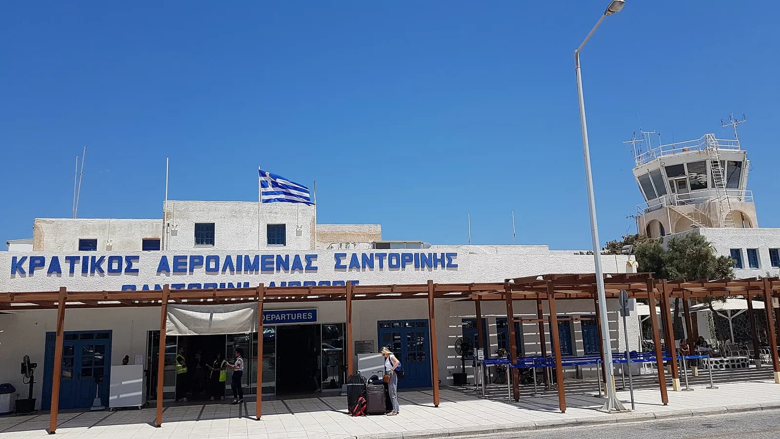 Santorini Airport landside terminal facade with DEPARTURES doors, control tower, and Greek flag