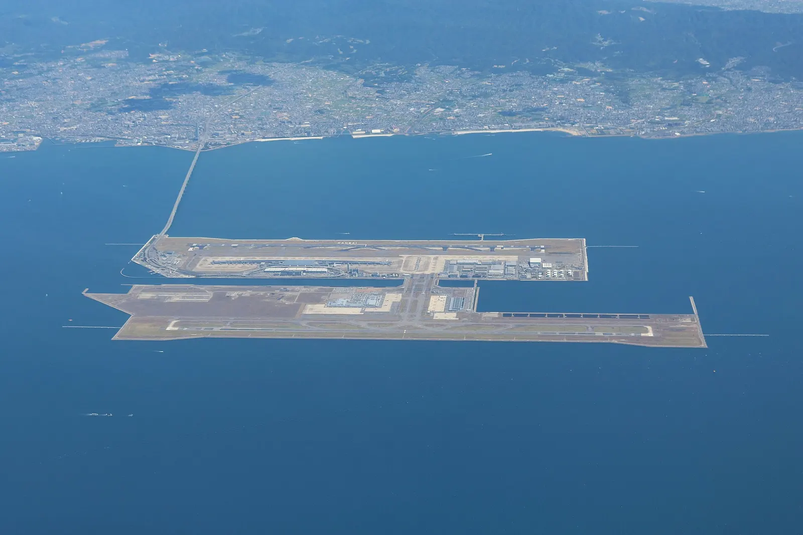 Wide aerial view of Kansai International Airport on an artificial island in Osaka Bay, with runways, terminal buildings, and surrounding water visible.