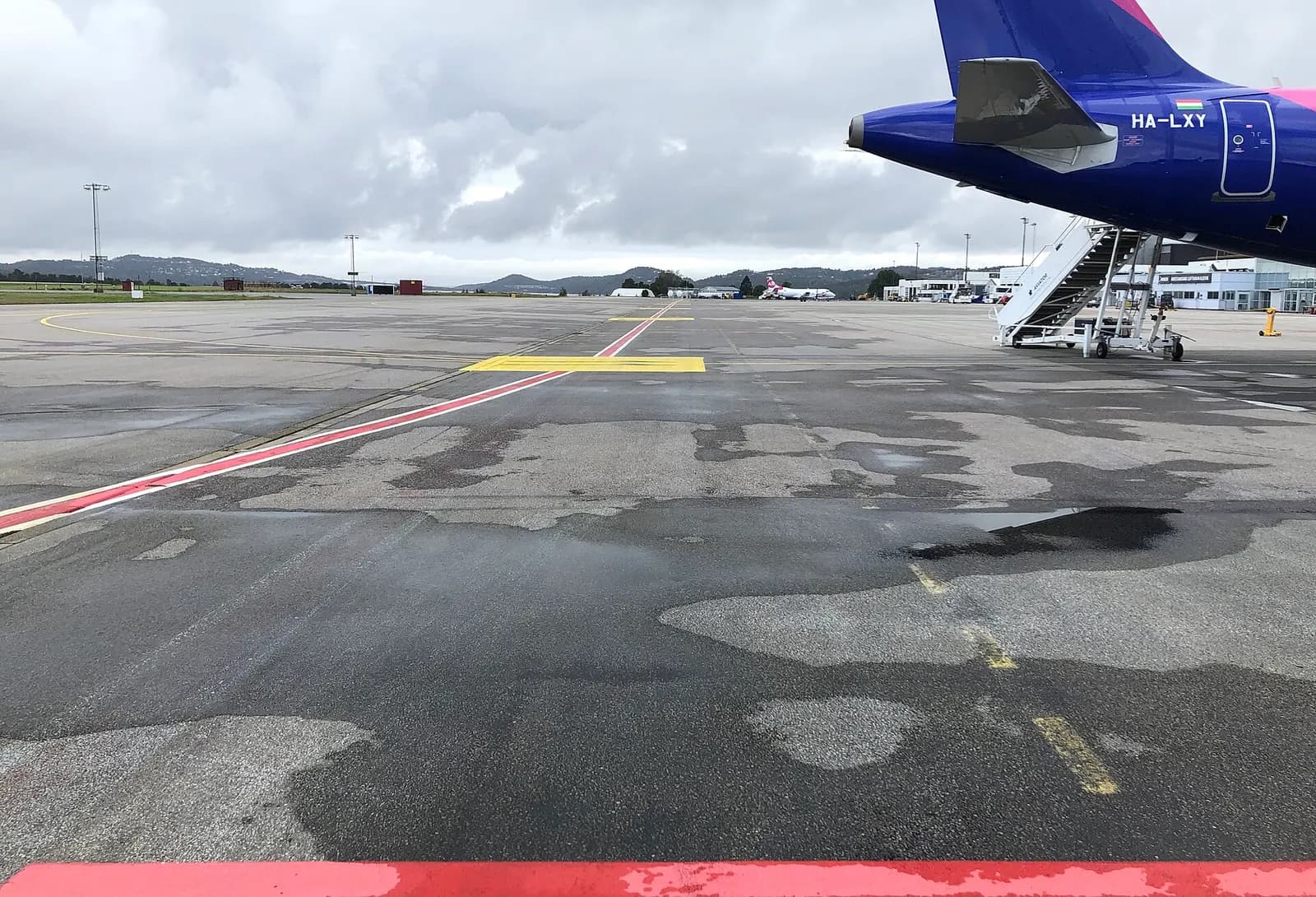 A view down the tarmac parking area from the northeasternmost stand 10 in direction of Kristiansand city. Two aircraft: Wizz Air A321 HA-LXY at stand 11 (arriving/departing GDN) and Sprint Air Saab 340 Freighter at 18 (arriving/departing CPH). Note the red line in the center/slightly left along TWY 