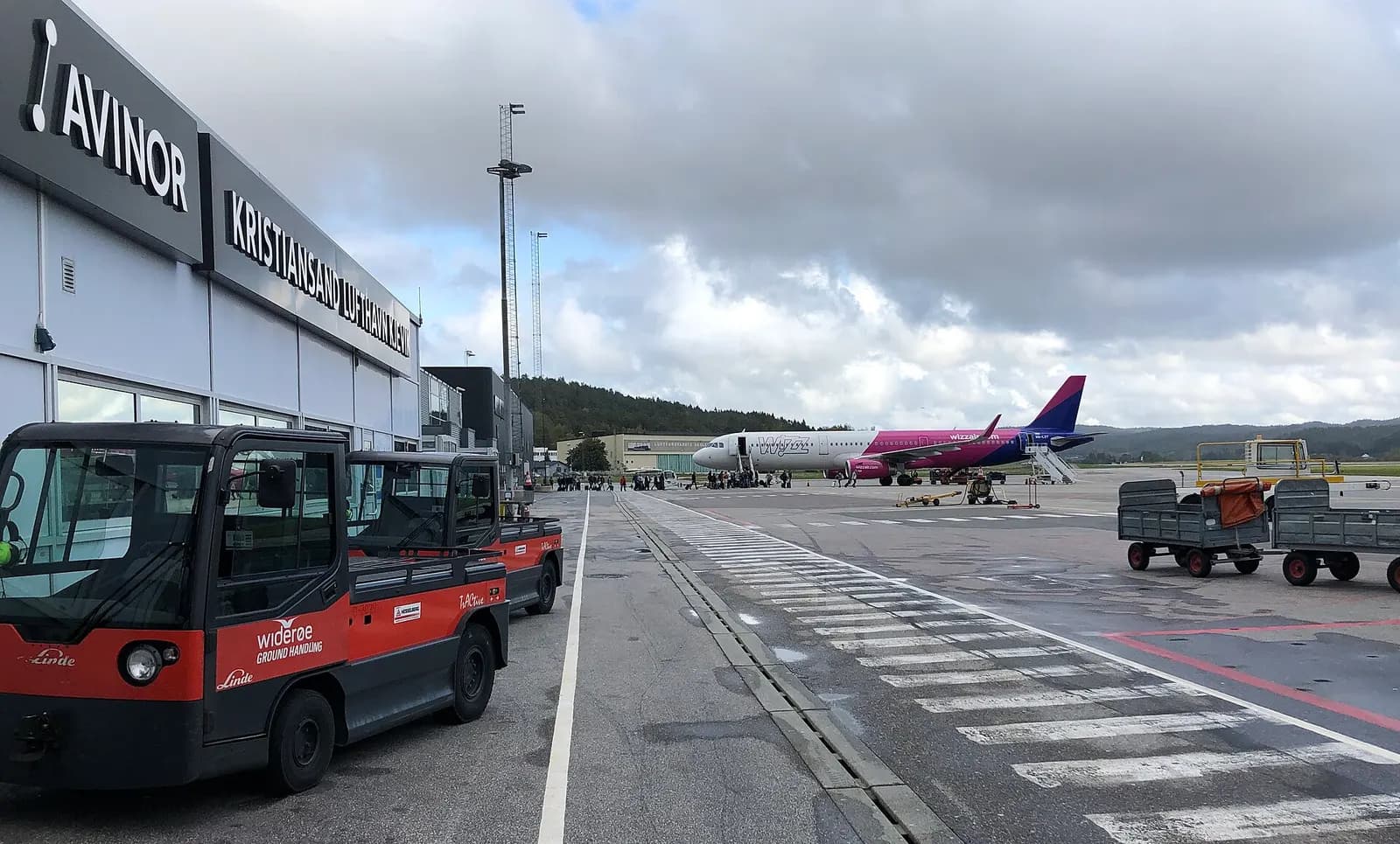 Overlooking the tarmac of Kristiansand Airport seen from the terminal access door for domestic arrivals. Several pieces of parked GSE of handling agents Wideroe WGH and Aviator, the latter serving Wizz Air A321 HA-LXY during boarding. Picture taken in late sep-2019