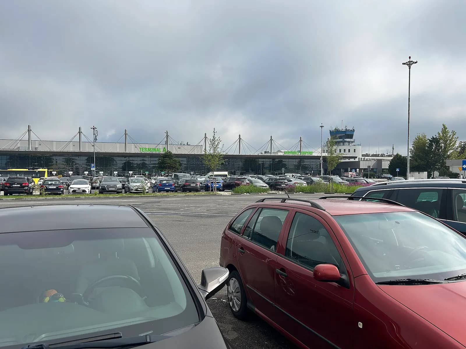 Rows of parked cars in the main outdoor parking lot in front of Katowice-Pyrzowice Airport on a summer day.