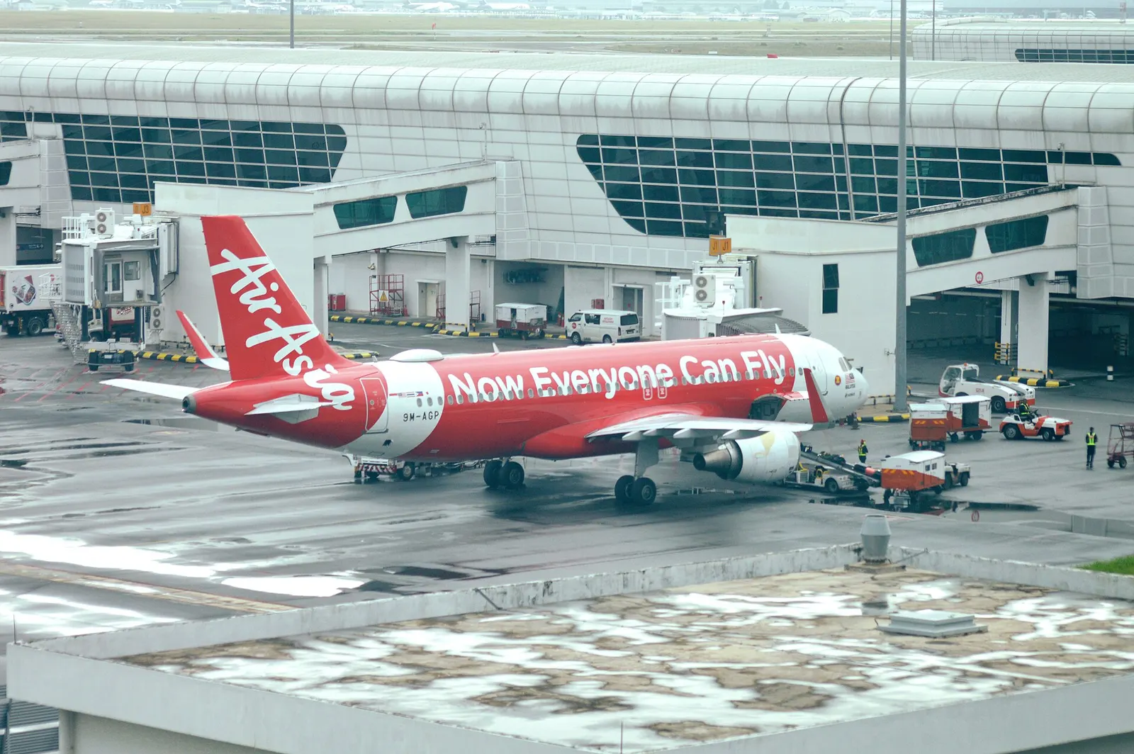 An AirAsia Airbus A320 is parked at a jet bridge gate with ground crew around it, viewed from inside Terminal 2.