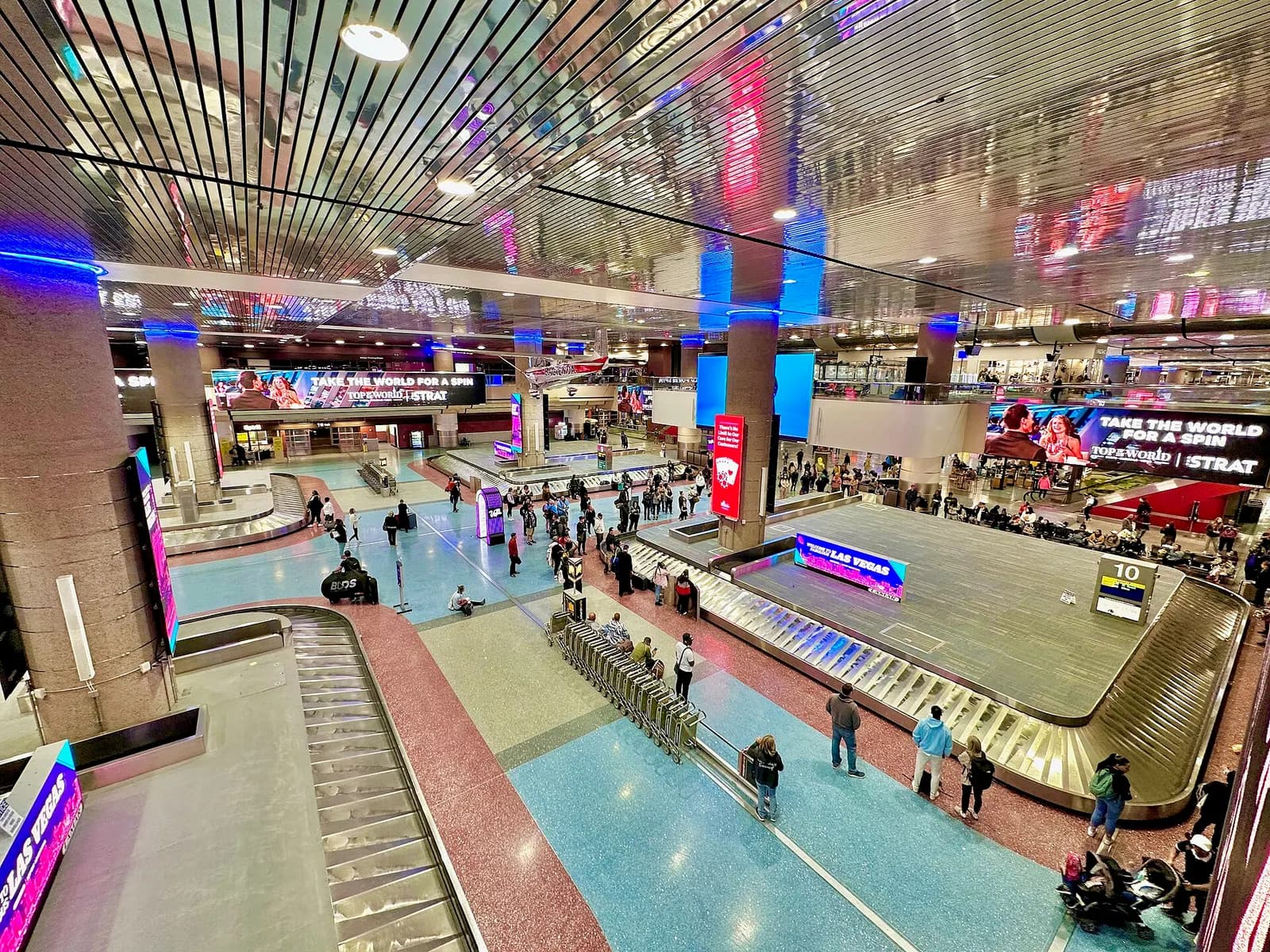 Baggage claim hall with multiple carousels, overhead flight information displays, and passengers collecting luggage.