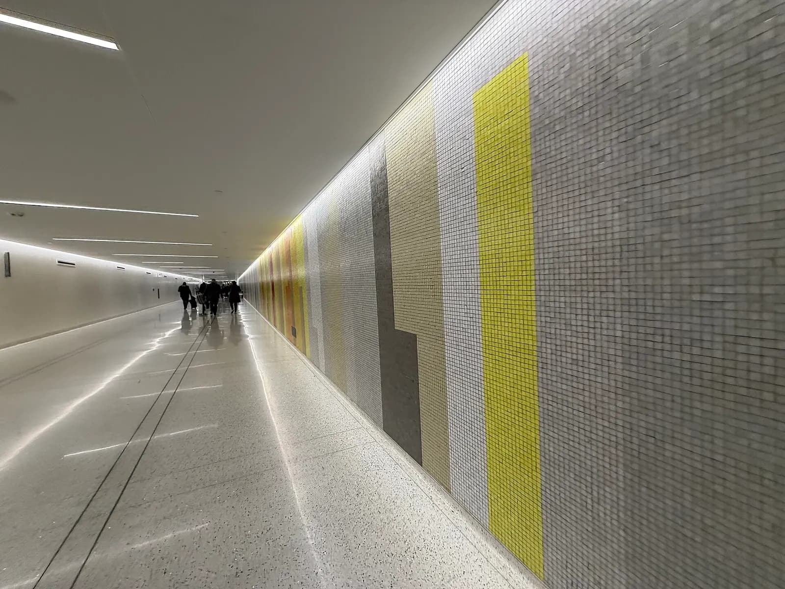 A long pedestrian tunnel in Terminal 4 lined with backlit panels in shifting rainbow colors, with travelers walking toward the gates.