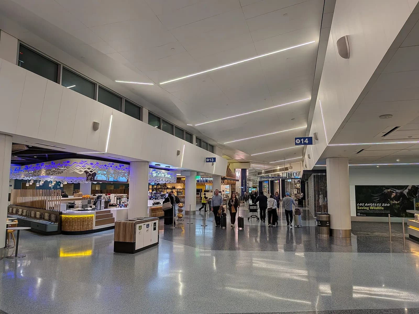 Passengers seated and walking through the gate holdroom in Terminal 1, with large windows overlooking the tarmac and parked aircraft.
