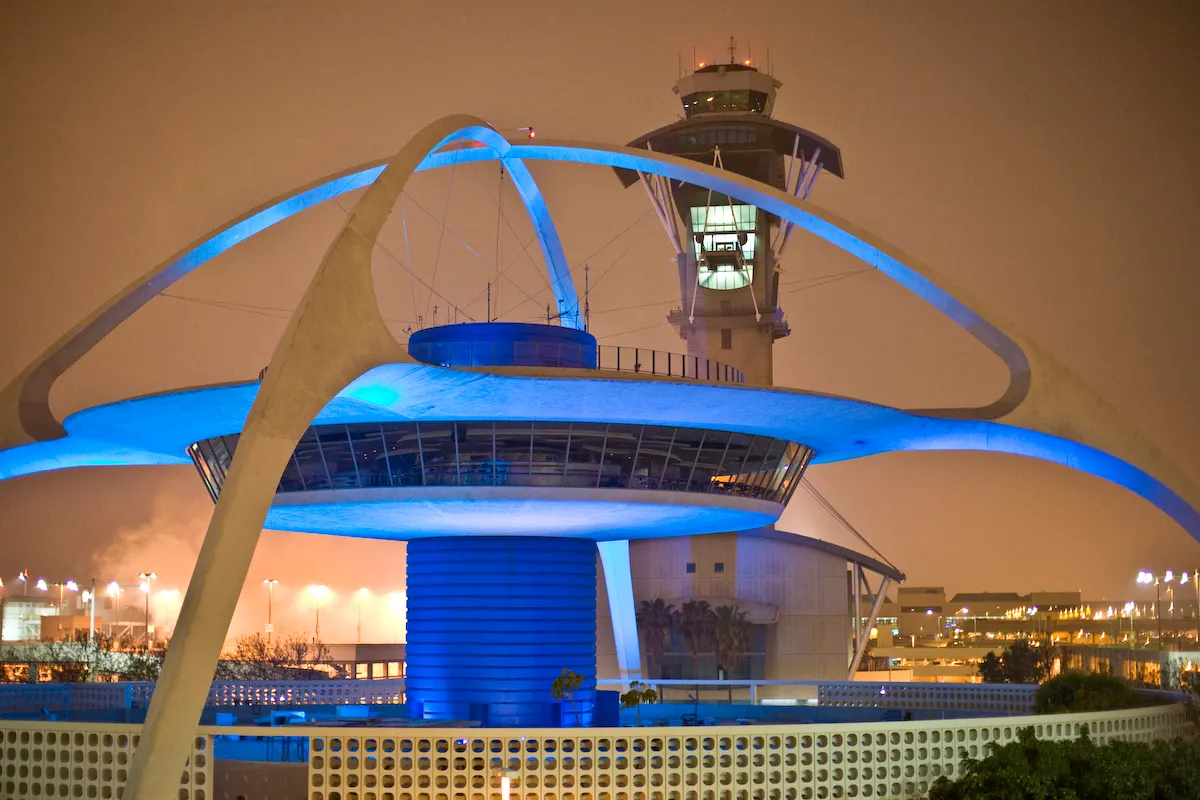 LAX's iconic mid-century Theme Building with its four crossed parabolic arches and the adjacent air traffic control tower against a clear sky.