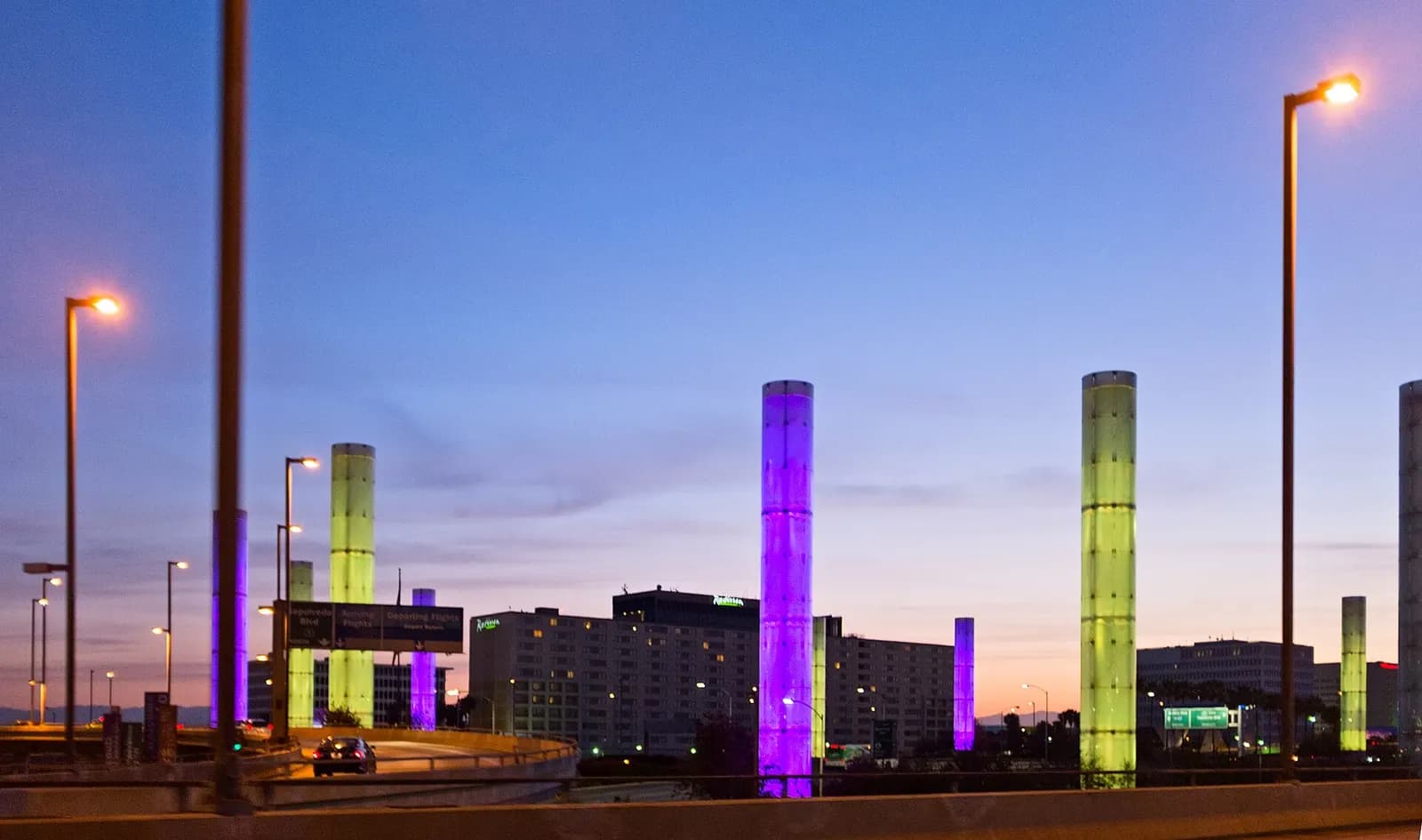Illuminated cylindrical light columns lining the highway approach to LAX in early morning light, with traffic lanes leading toward the airport.