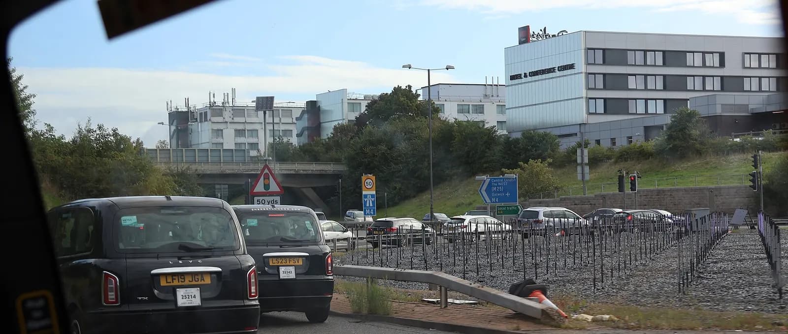 The main road entrance to London Heathrow Airport with terminal buildings, landscaped medians, and vehicles approaching drop-off areas.