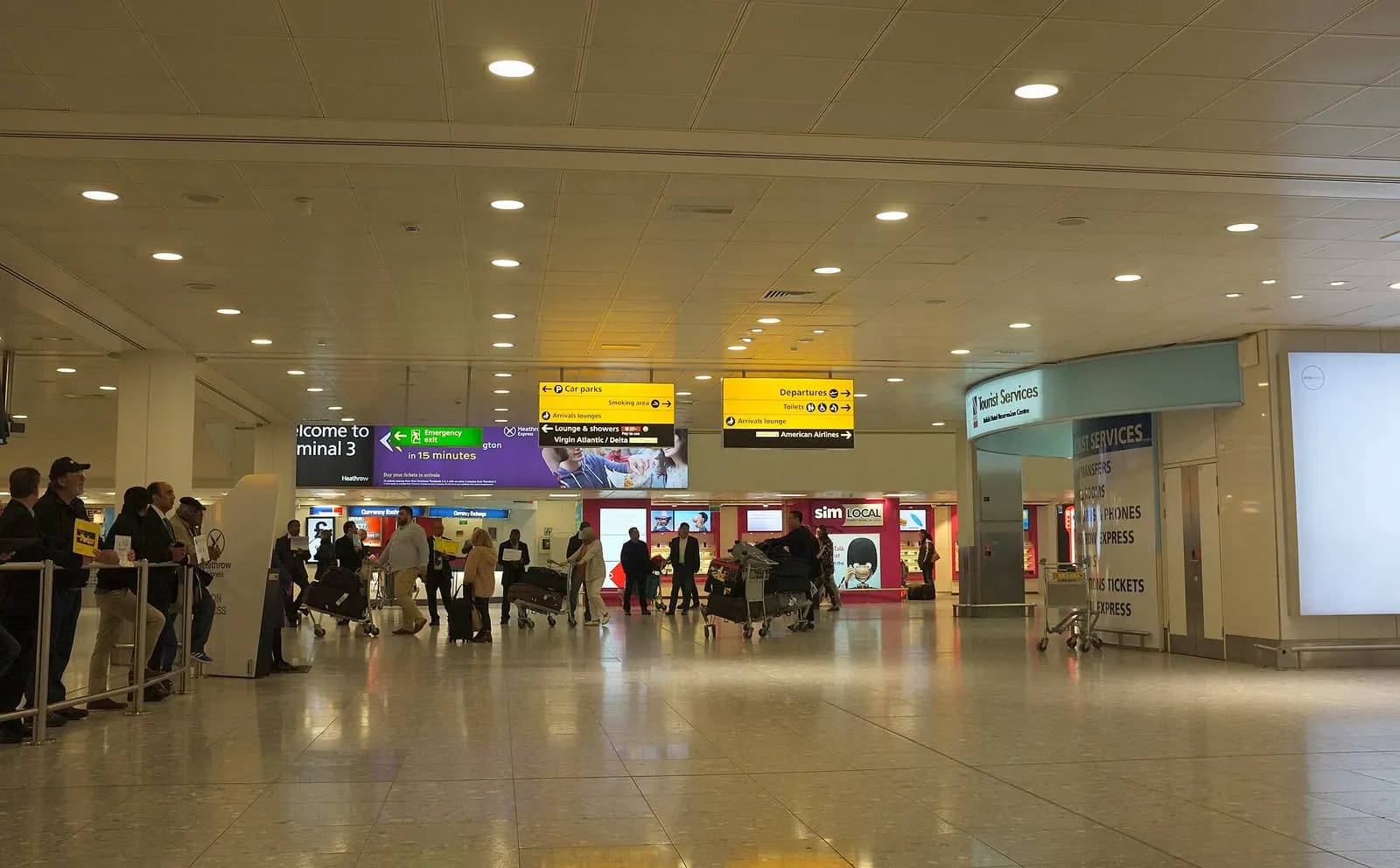 The arrivals hall inside Heathrow Terminal 3 with baggage carousel areas, directional signs, and passengers exiting through glass doors.