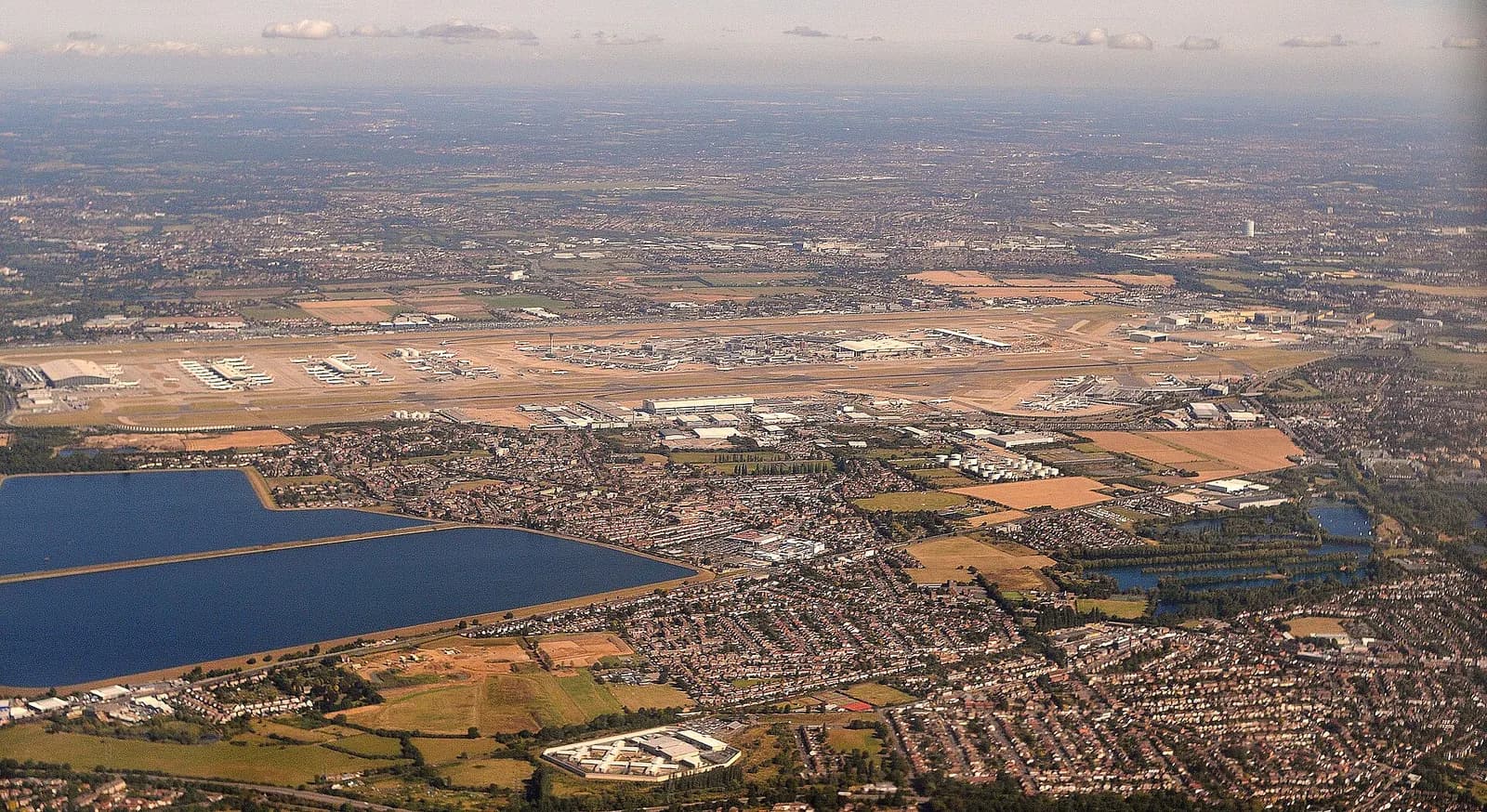 A broad landscape view of London Heathrow Airport showing multiple runways, terminal buildings, and an aircraft on approach in the distance.