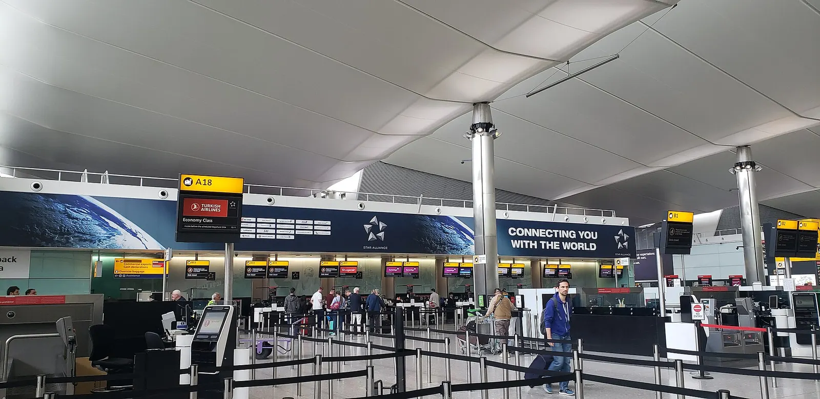 A wide, bright view of the London Heathrow Terminal 2 departures hall with check-in counters, departure screens, and travelers moving through the open concourse.