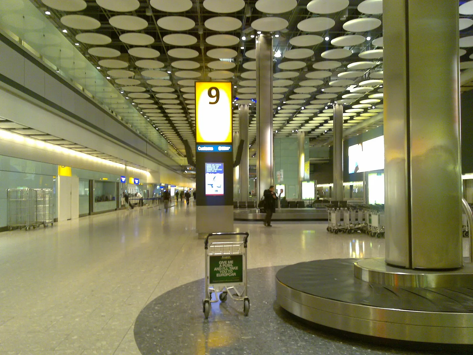 The baggage reclaim area in Heathrow Terminal 5 with multiple carousels, overhead flight information displays, and travelers collecting luggage.