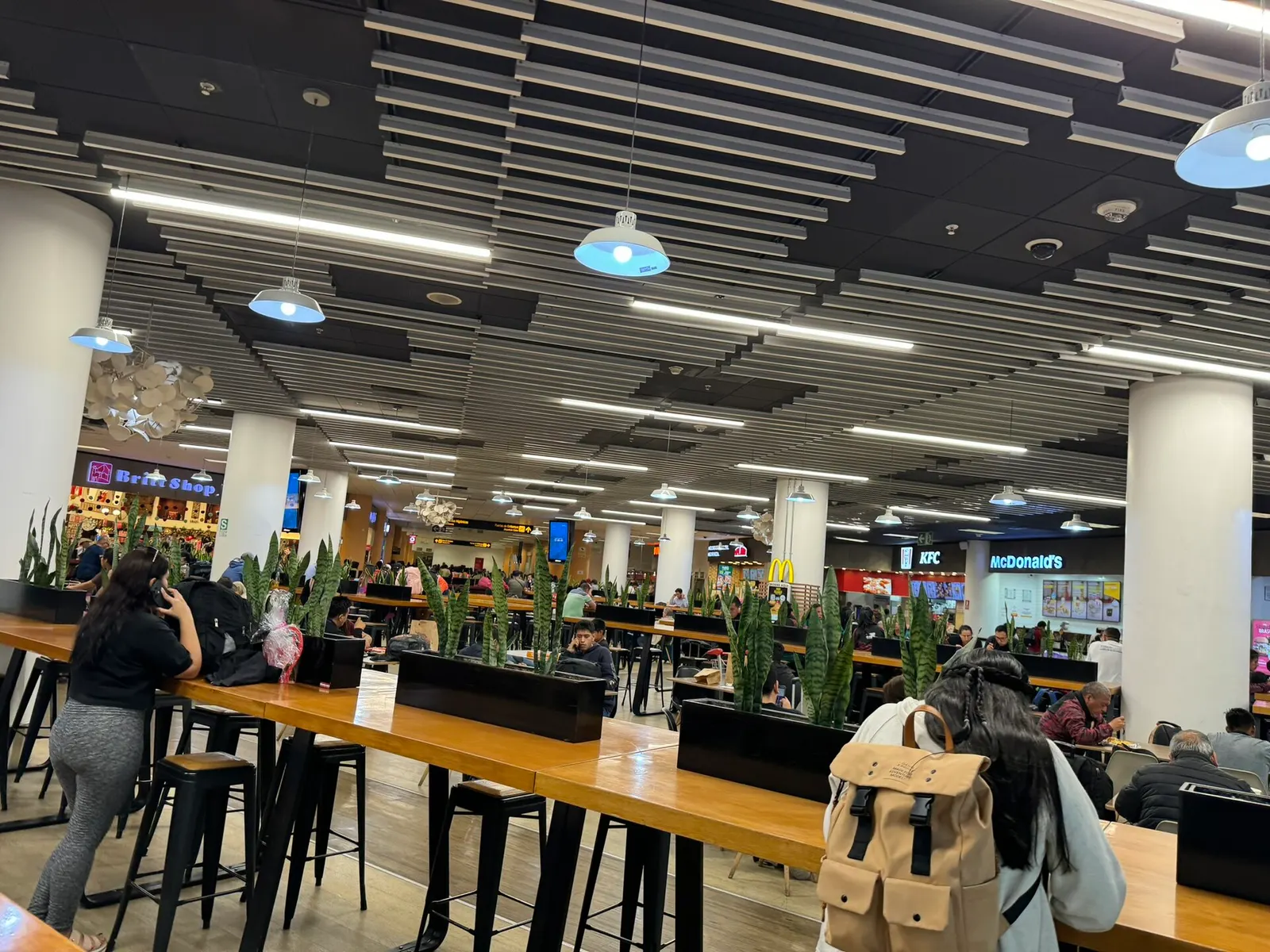 An airport food court with restaurant counters, dining tables, and passengers eating in the terminal concourse.
