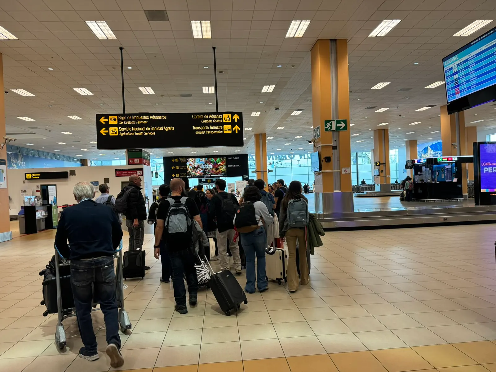 International baggage claim hall with multiple carousels, overhead flight information displays, and arriving passengers.