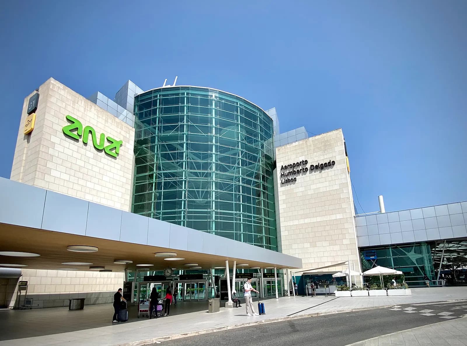 The glass-fronted main terminal building of Humberto Delgado Airport seen from the approach road on a sunny day.
