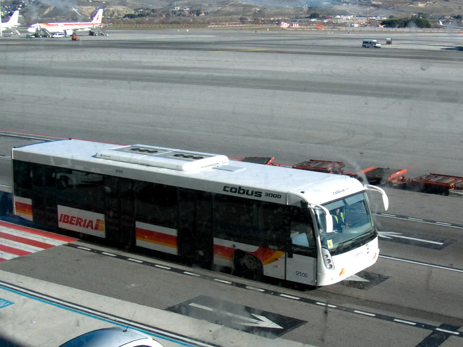 A white Iberia Cobus 3000 passenger shuttle bus on the ramp, used to ferry travelers between terminals and remote stands.